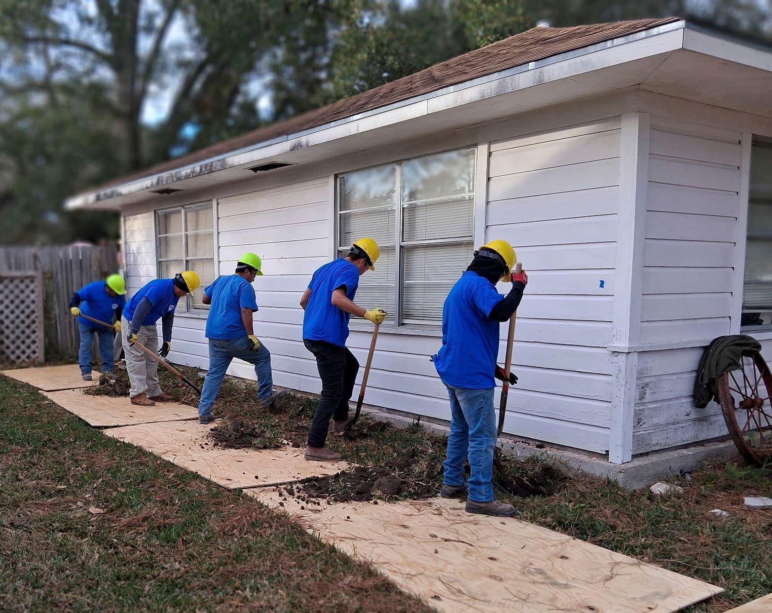 Five Allied Foundation workers in blue shirts and yellow helmets digging along the side of a white house