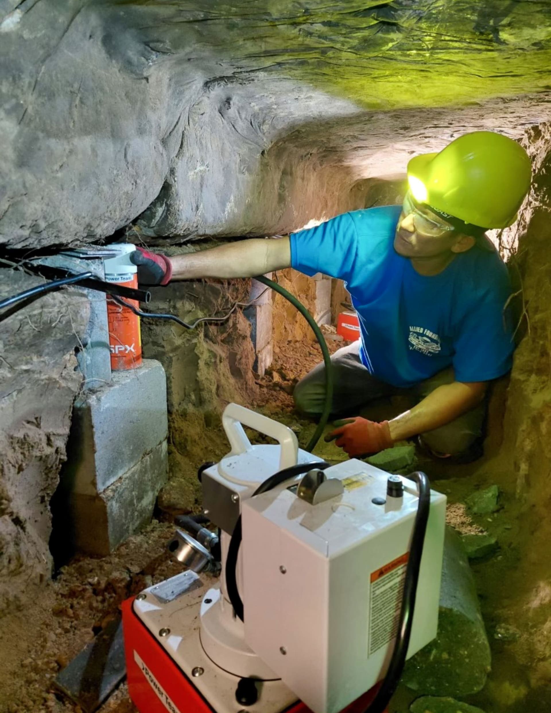 Allied Foundation worker in a yellow helmet and blue shirt operating a ramming machine inside an access tunnel beneath a home