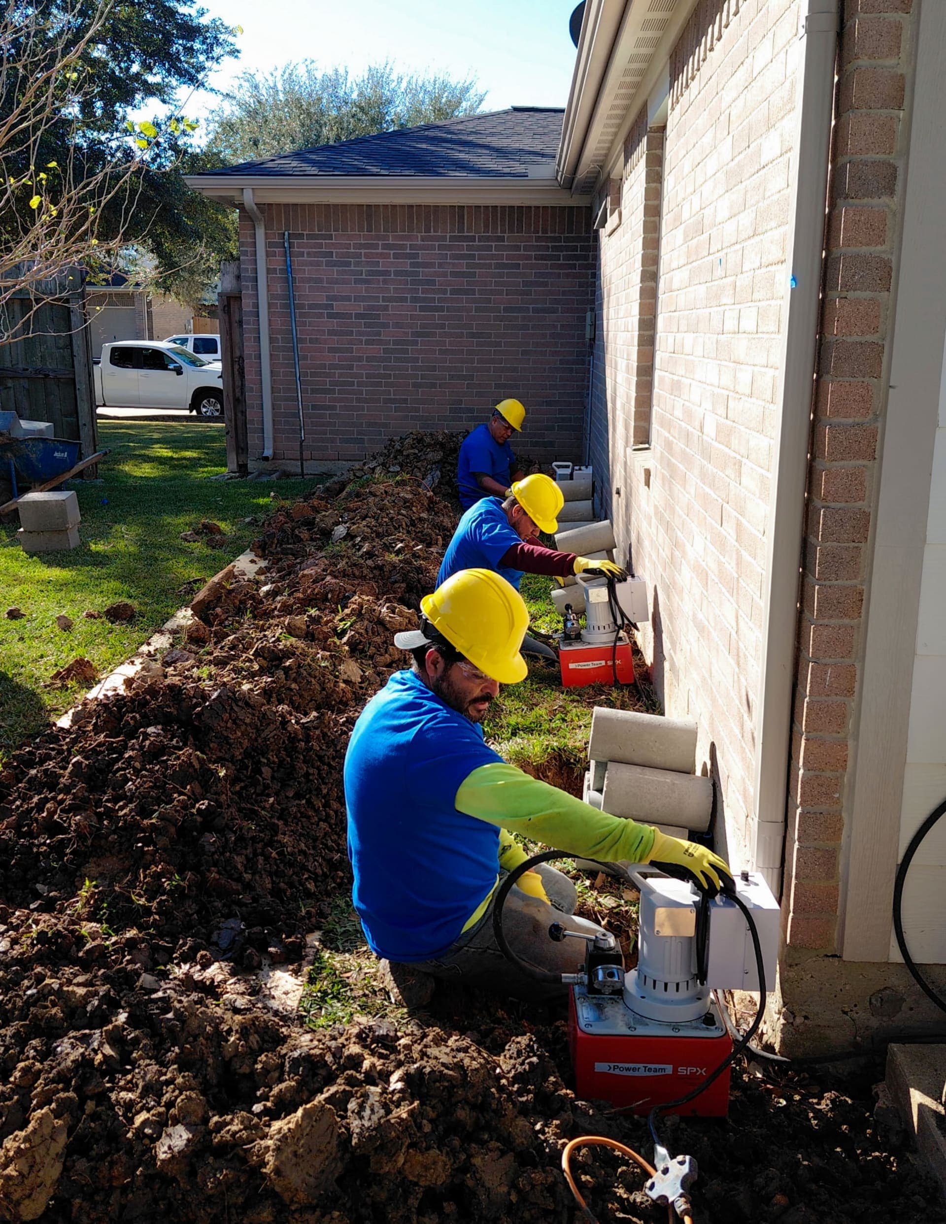 Three Allied Foundation workers in blue shirts and yellow helmets ramming piers along the left side of a brick wall